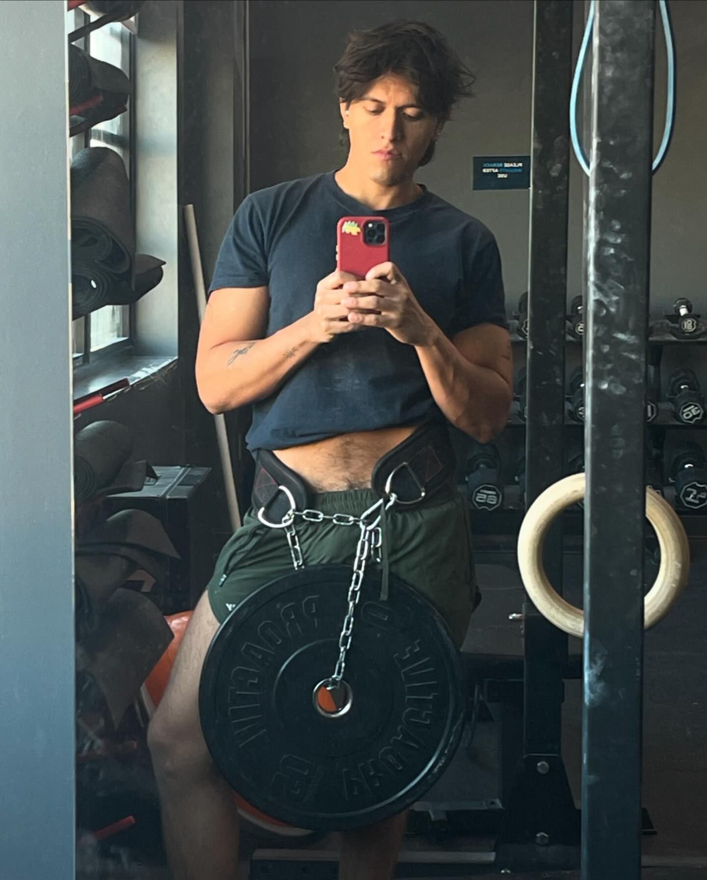 Fit man taking a gym selfie with a weight plate suspended by a chain from his weightlifting belt, focusing on strength and fitness.