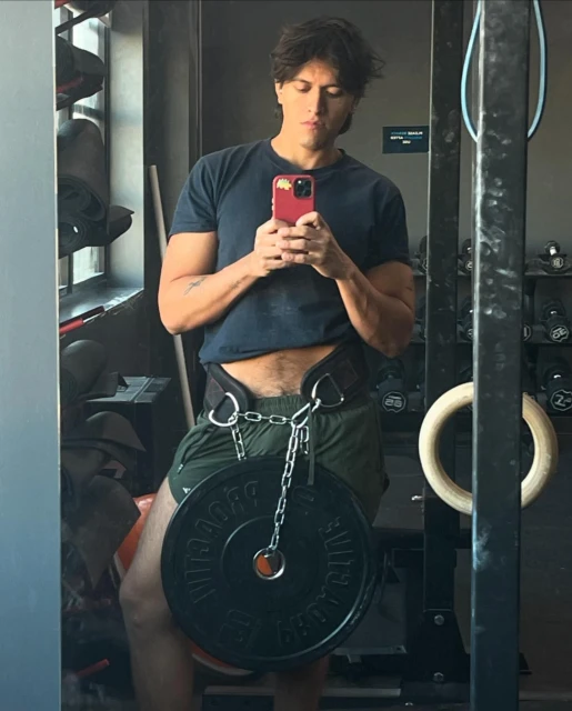 Fit man taking a gym selfie with a weight plate suspended by a chain from his weightlifting belt, focusing on strength and fitness.