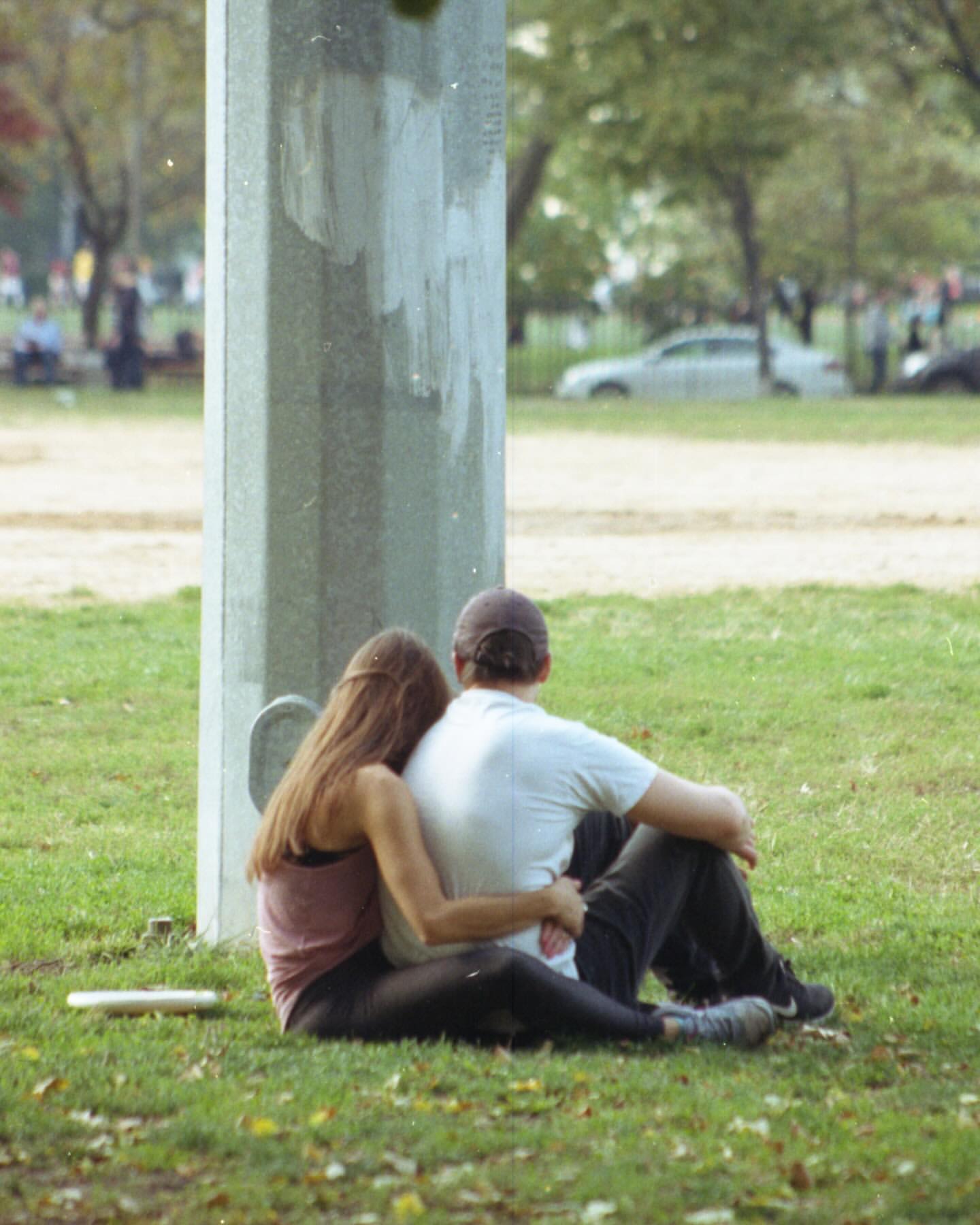 Couple sitting on grass in park next to concrete structure, embraced in daylight with soft focus. Trees and car in distance.