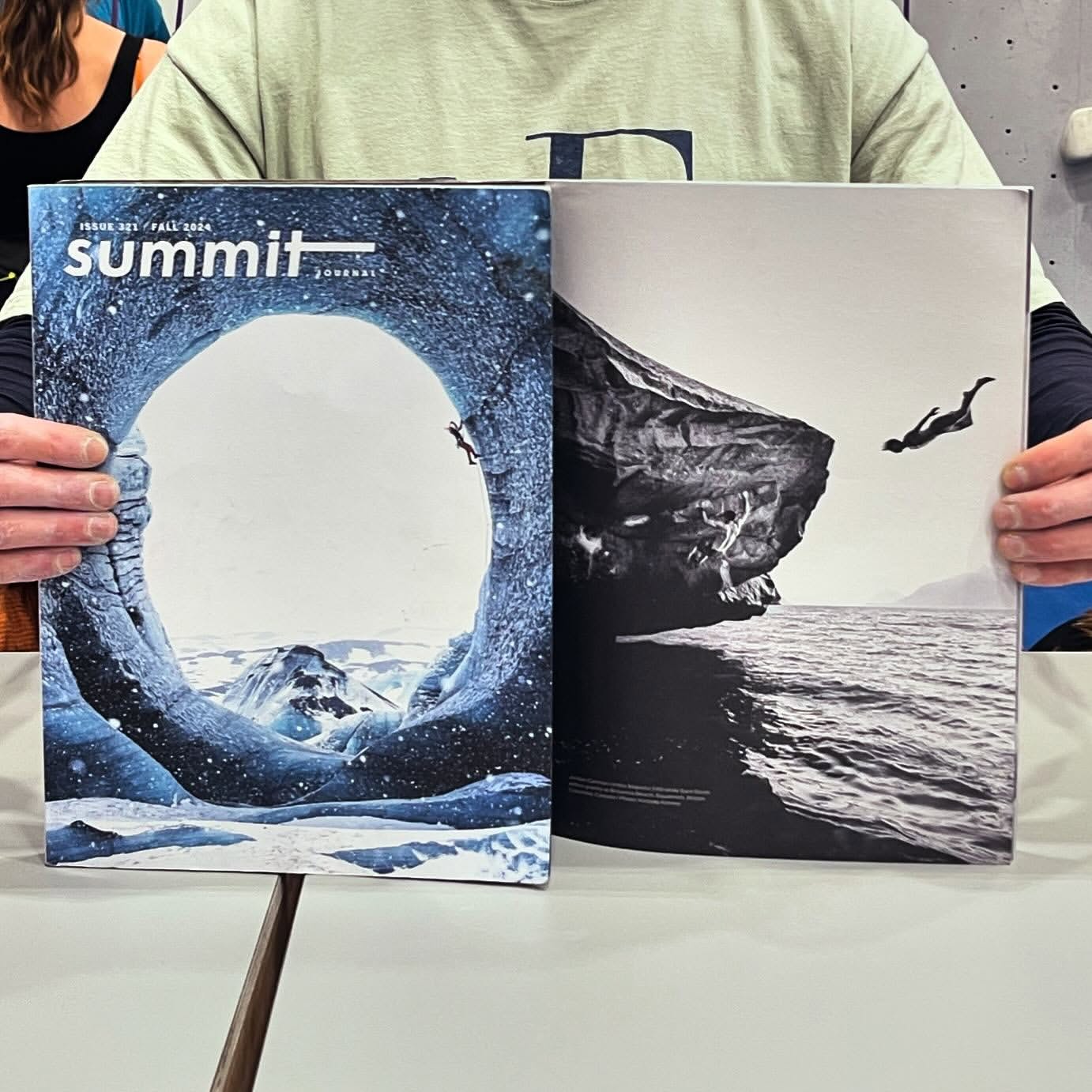 Two magazines featuring nature and adventure photos are held up, including a climber in an ice cave and a cliff diver above the sea.