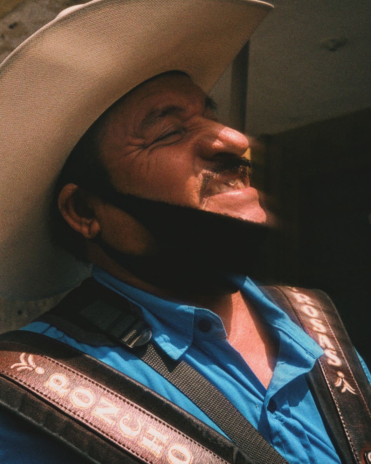 Smiling mature man wearing a cowboy hat, blue shirt, and leather guitar strap in a sunlit outdoor setting.