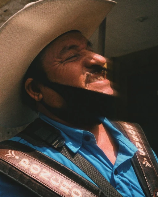 Smiling mature man wearing a cowboy hat, blue shirt, and leather guitar strap in a sunlit outdoor setting.