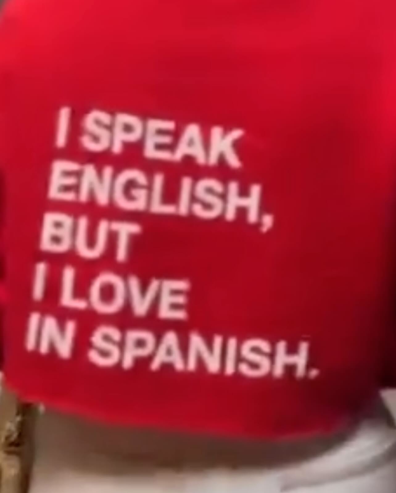 Close-up of a red sweatshirt with the phrase 'I SPEAK ENGLISH, BUT I LOVE IN SPANISH' in white sans-serif typeface on the chest.