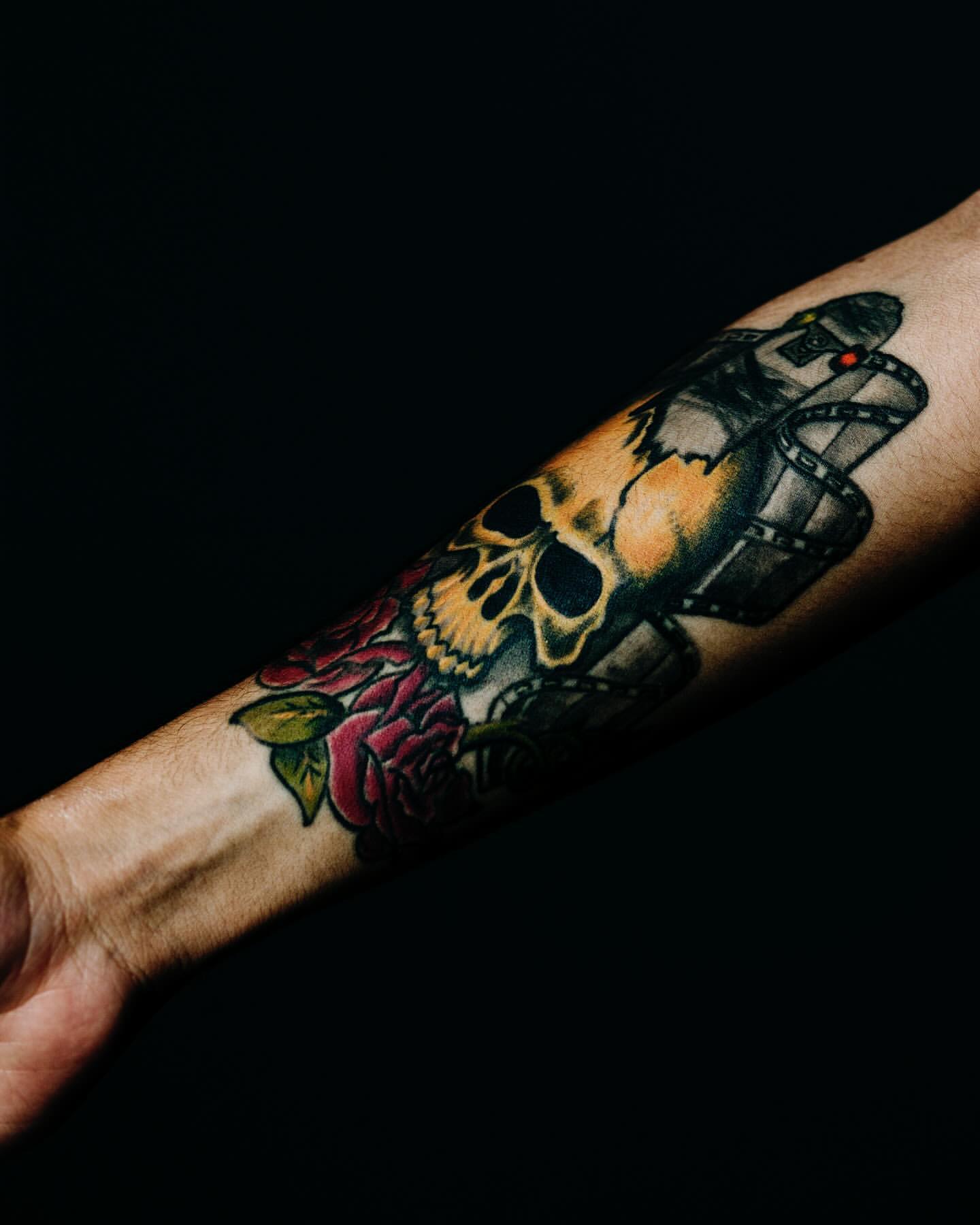 Arm tattoo of a skull, film reel, and roses, displaying gothic art and dark themes against a solid black background in studio lighting.