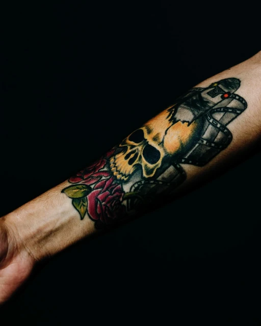 Arm tattoo of a skull, film reel, and roses, displaying gothic art and dark themes against a solid black background in studio lighting.