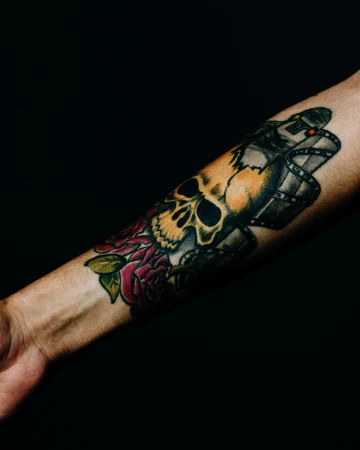 Arm tattoo of a skull, film reel, and roses, displaying gothic art and dark themes against a solid black background in studio lighting.