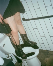 Close-up of a person wearing black cowboy boots standing on a white toilet in a tiled restroom, photographed by Andres Burgos.