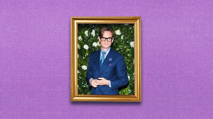 Portrait of a man in a blue suit and glasses, framed by a gold frame and a green and white flower background, on a purple textured backdrop.