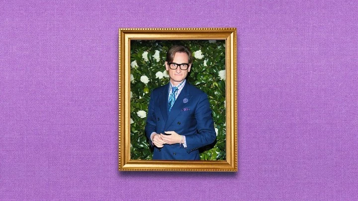 Portrait of a man in a blue suit and glasses, framed by a gold frame and a green and white flower background, on a purple textured backdrop.