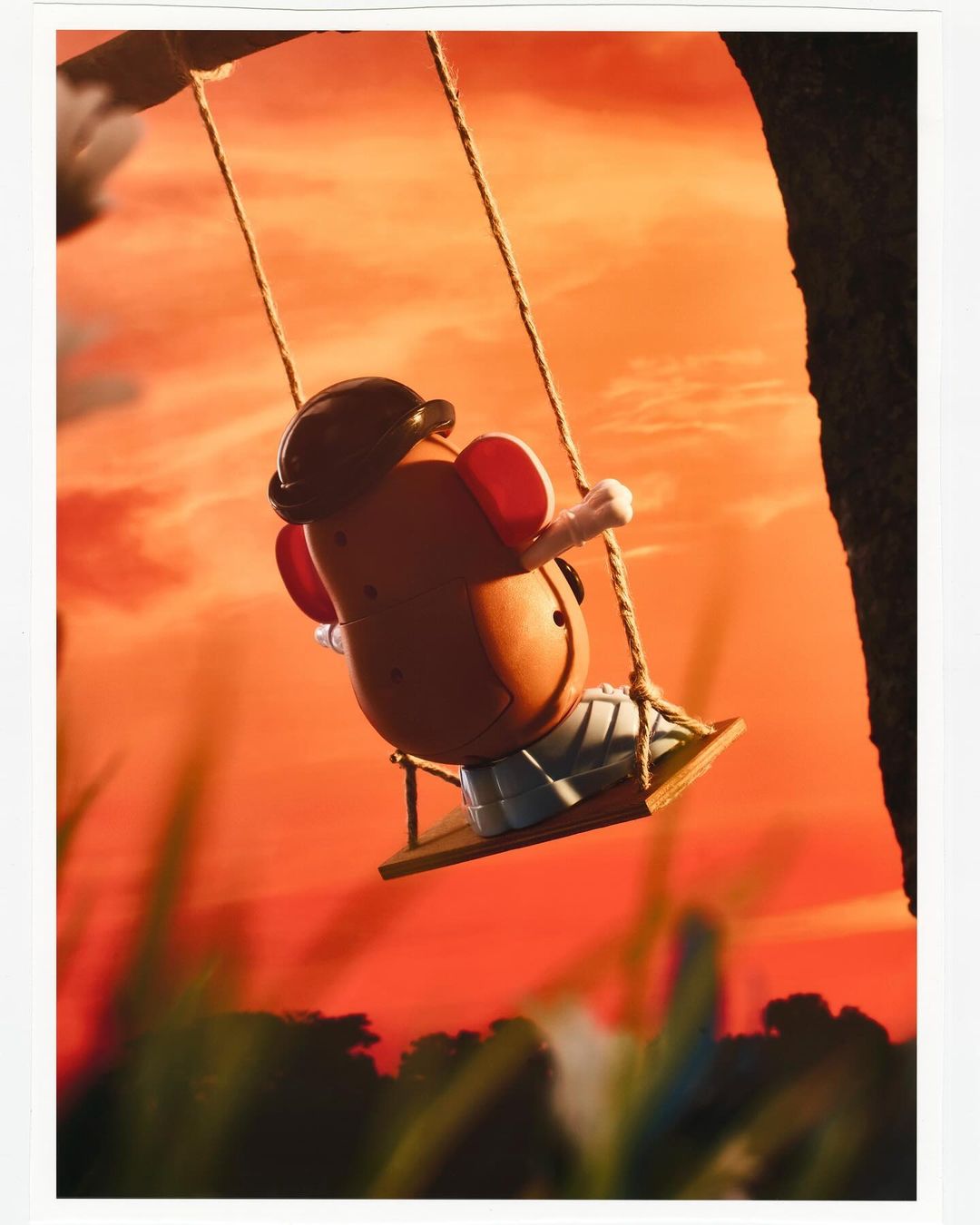 Toy Mr. Potato Head sits on a wooden swing hanging from a tree against a vibrant orange sunset background in a captivating image.