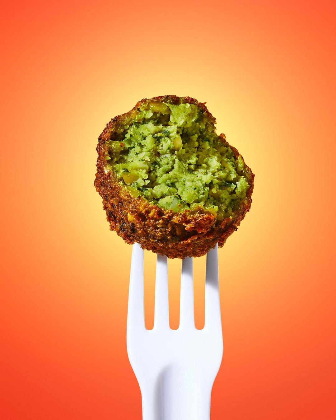 A single falafel ball on a white plastic fork, brightly lit against an orange gradient backdrop for a food presentation.