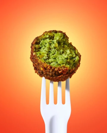 A single falafel ball on a white plastic fork, brightly lit against an orange gradient backdrop for a food presentation.