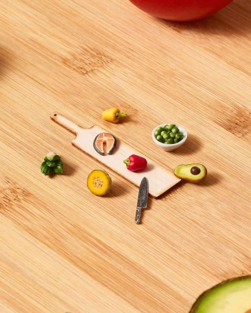 Miniature food art on a wooden cutting board featuring vegetables, salmon, and a knife, all arranged on a wood background.