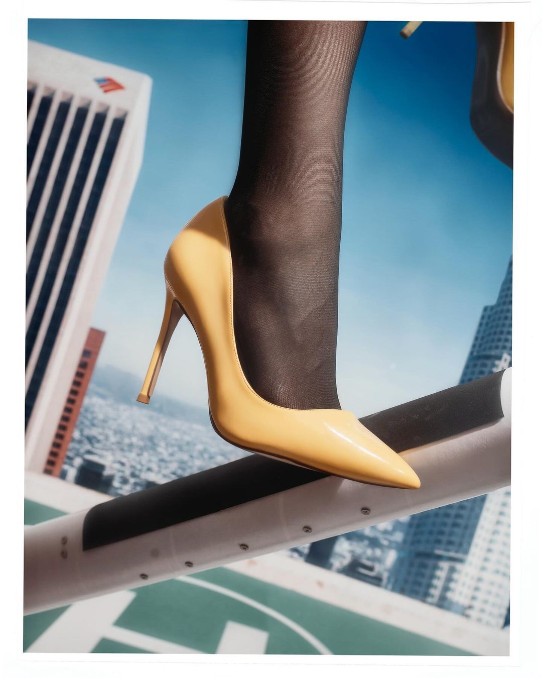 Woman's leg wearing sheer black stockings and a beige high heel pump, on an elevated walkway, with a city skyline in the background.