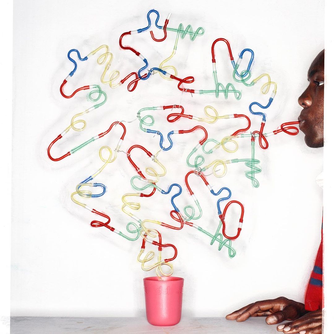 African American male blowing air through a red drinking straw connected to a cluster of colorful, looped straws over a pink cup.