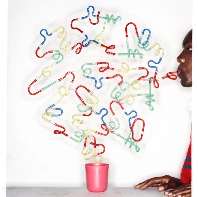 African American male blowing air through a red drinking straw connected to a cluster of colorful, looped straws over a pink cup.