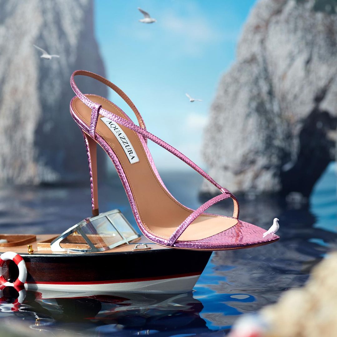 Elevated pink strappy Aquazzura stiletto on a miniature boat in water with coastal cliffs in the background and seagulls flying overhead.