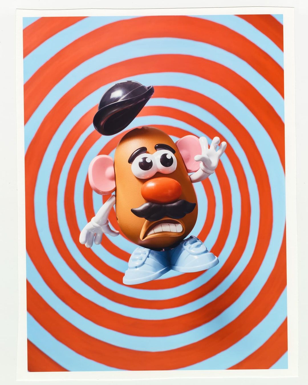 Toy potato figure with hat floating above its head, posed in front of a concentric circle background, art photography.