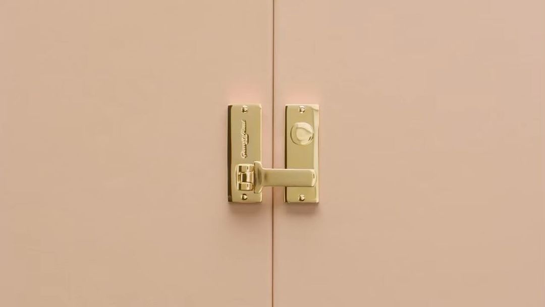 Close-up of a gold door handle and lock on a light pink, double-door cabinet or closet, creating a minimalist, elegant, modern aesthetic.
