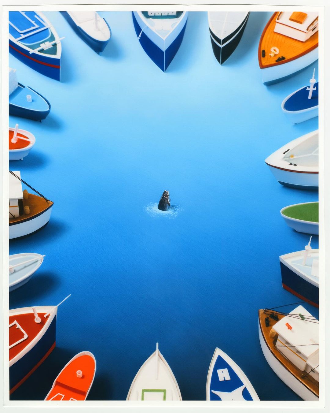 A miniature seal swims in blue water, surrounded by a circle of colorful toy boats in a high-angle, studio-lit composition.