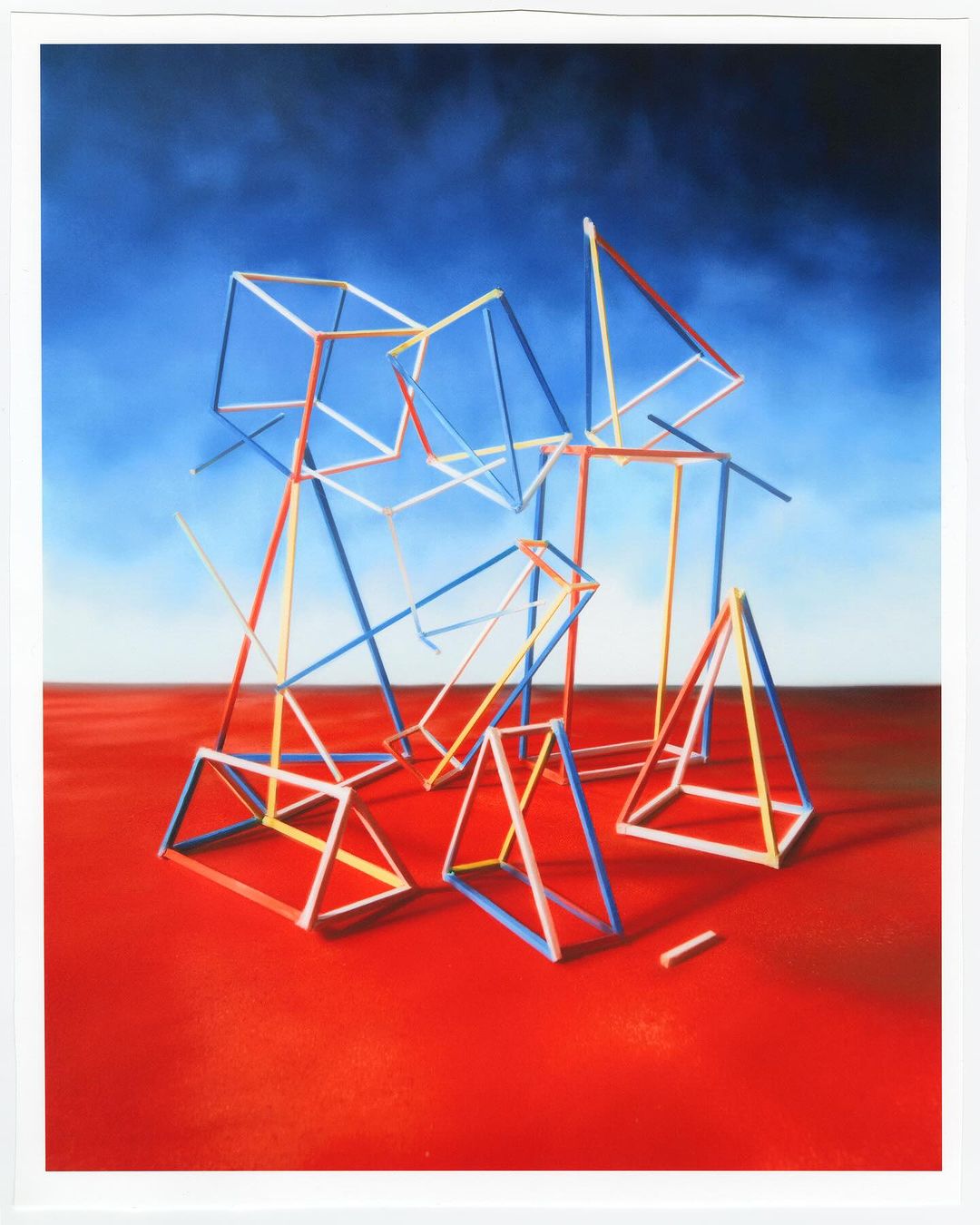 Abstract geometric art piece with multi-colored straw-like structures against a blue sky and a red-colored surface creating a modern, surreal scene.