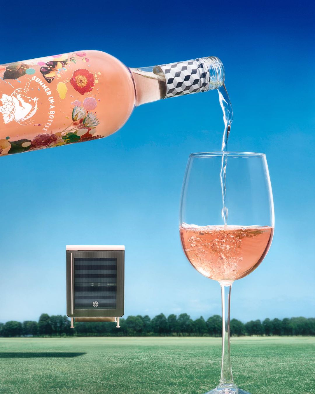 Rose wine being poured into a glass, set against a blue sky and green grass, alongside a small wine refrigerator, creating a surreal outdoor wine scene.
