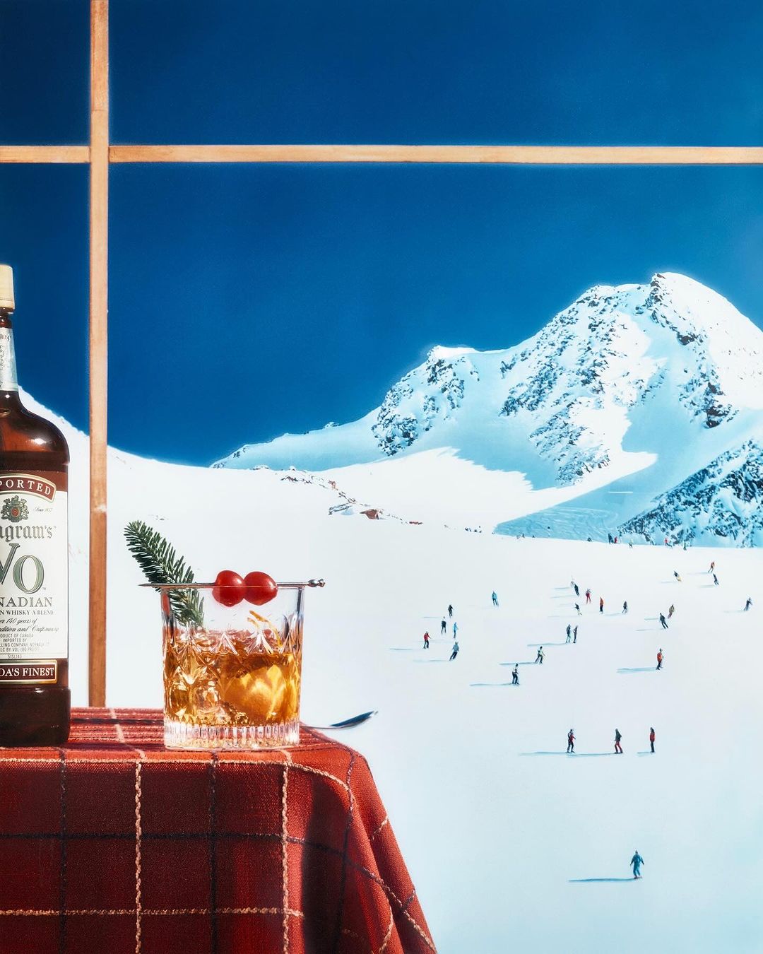 Cocktail and liquor bottle on a red tablecloth, snowy mountains and blue sky background, skiers visible in the distance.