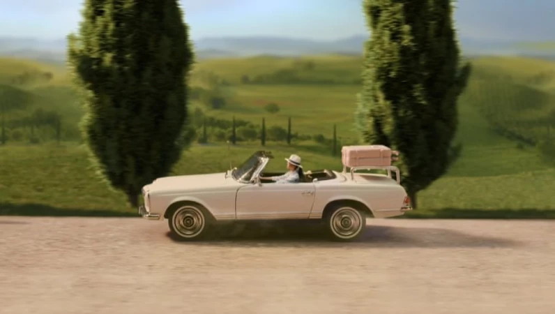 Woman driving a vintage white convertible car with a pink suitcase on a rack, travelling through a green, rural landscape.