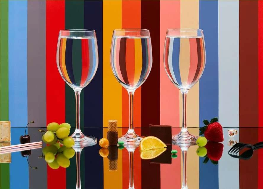 Three glasses of water reflect colorful stripes, accompanied by grapes, lemon, cherry, strawberry and sweets on a glossy black surface.