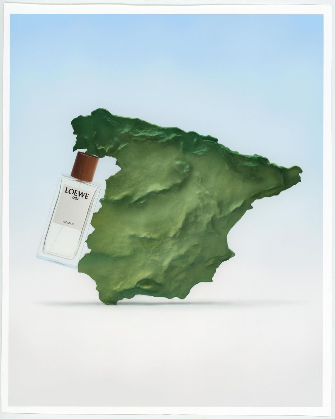 Still life of a Loewe perfume bottle next to a green textured landmass under bright lighting and pale blue gradient background.