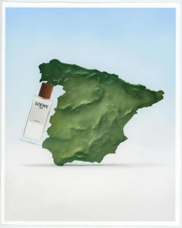 Still life of a Loewe perfume bottle next to a green textured landmass under bright lighting and pale blue gradient background.