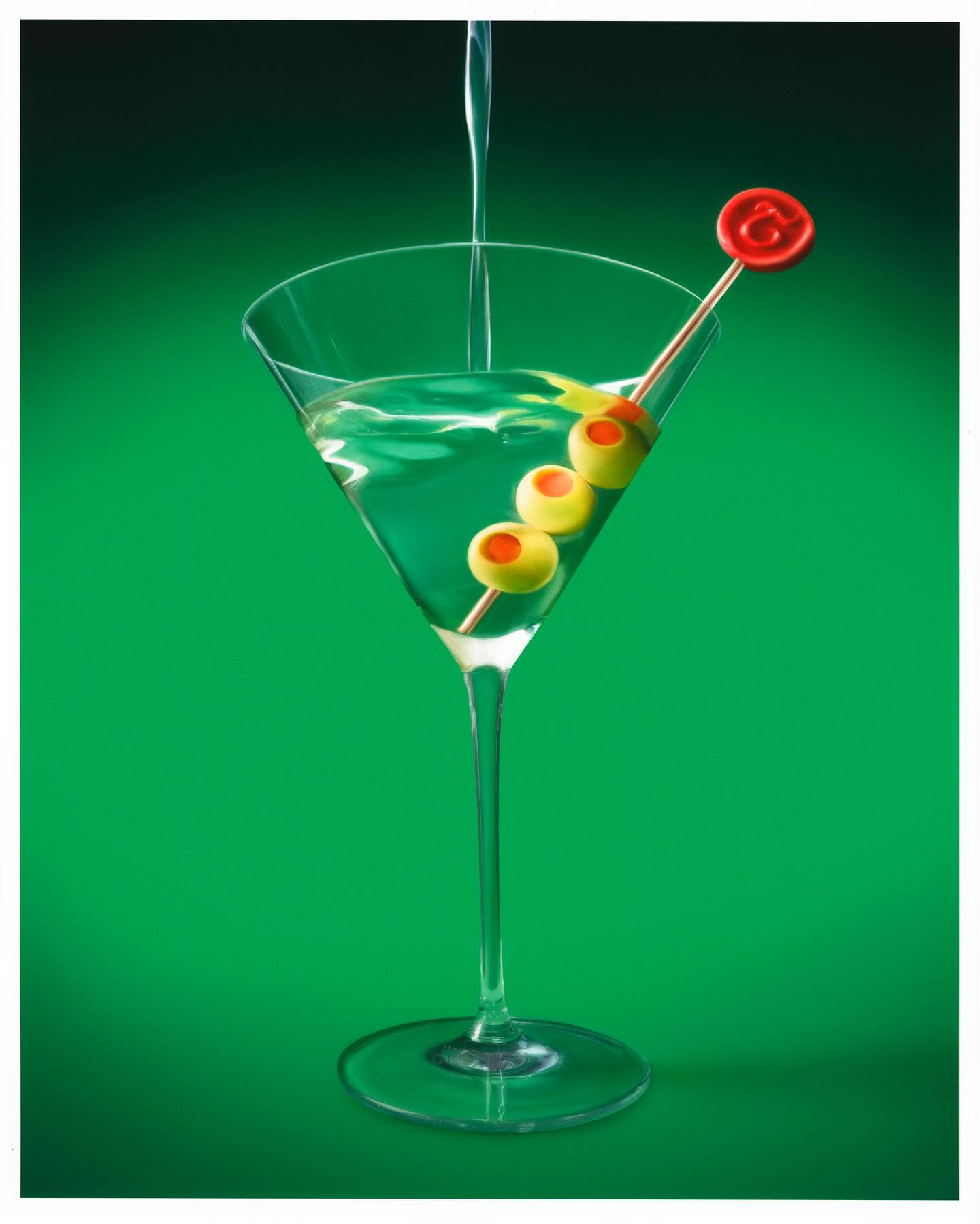 Martini glass being filled with liquid. Green olives on a toothpick garnish with a red cap, against a graduated green background.