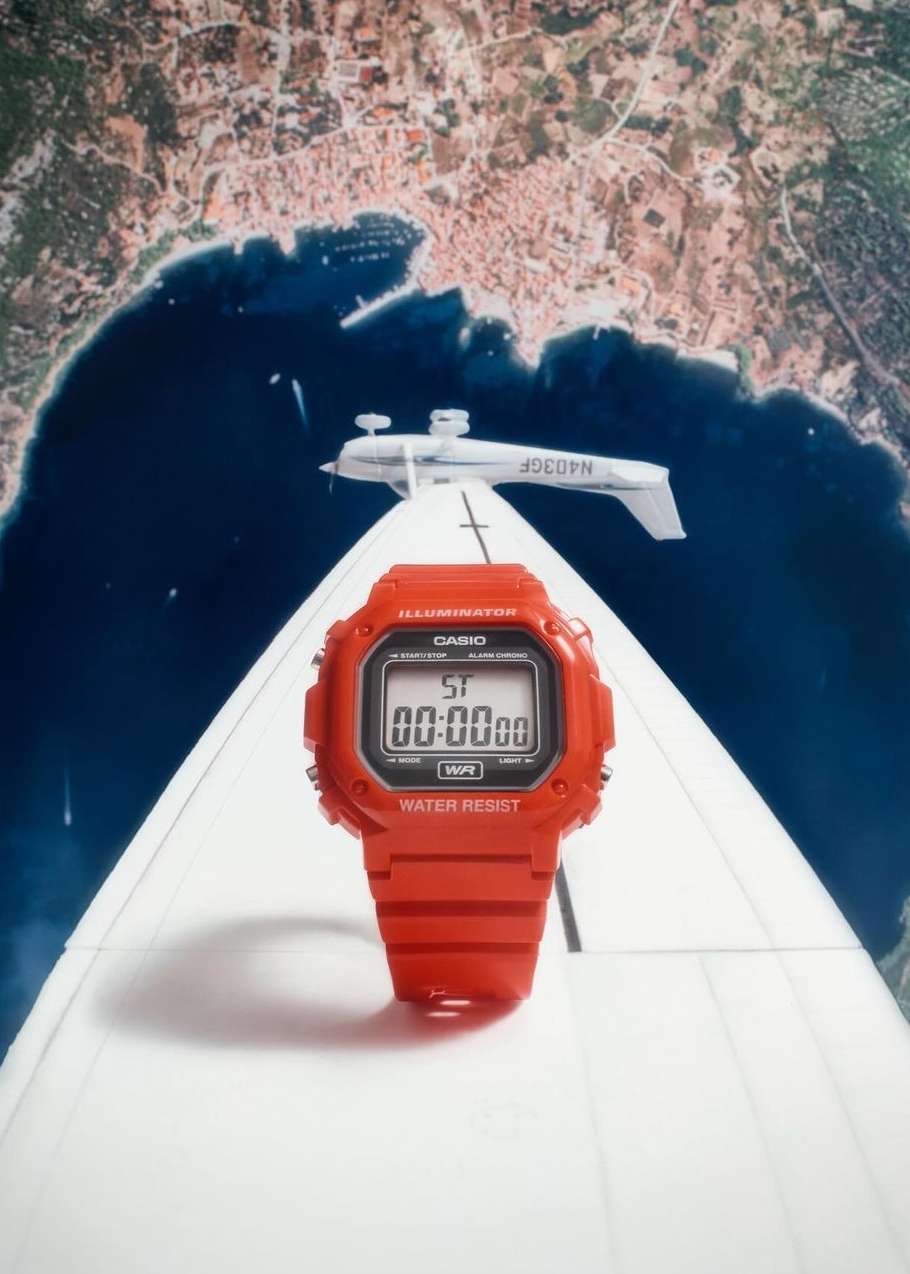 Red digital watch stands upright on a white airplane wing against a backdrop of a coastal city and dark blue water on a bright day.