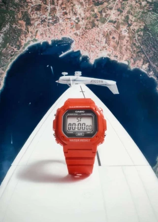 Red digital watch stands upright on a white airplane wing against a backdrop of a coastal city and dark blue water on a bright day.