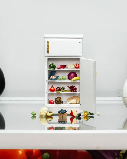 A miniature white refrigerator with a variety of colorful miniature fruits, vegetables, and eggs inside, set against a white background.