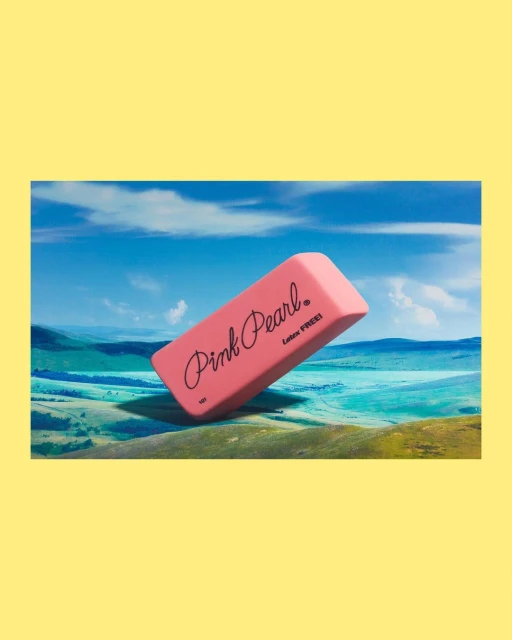Pink Pearl eraser floating above a green and blue landscape under a bright blue sky, creating a surreal and playful composition.