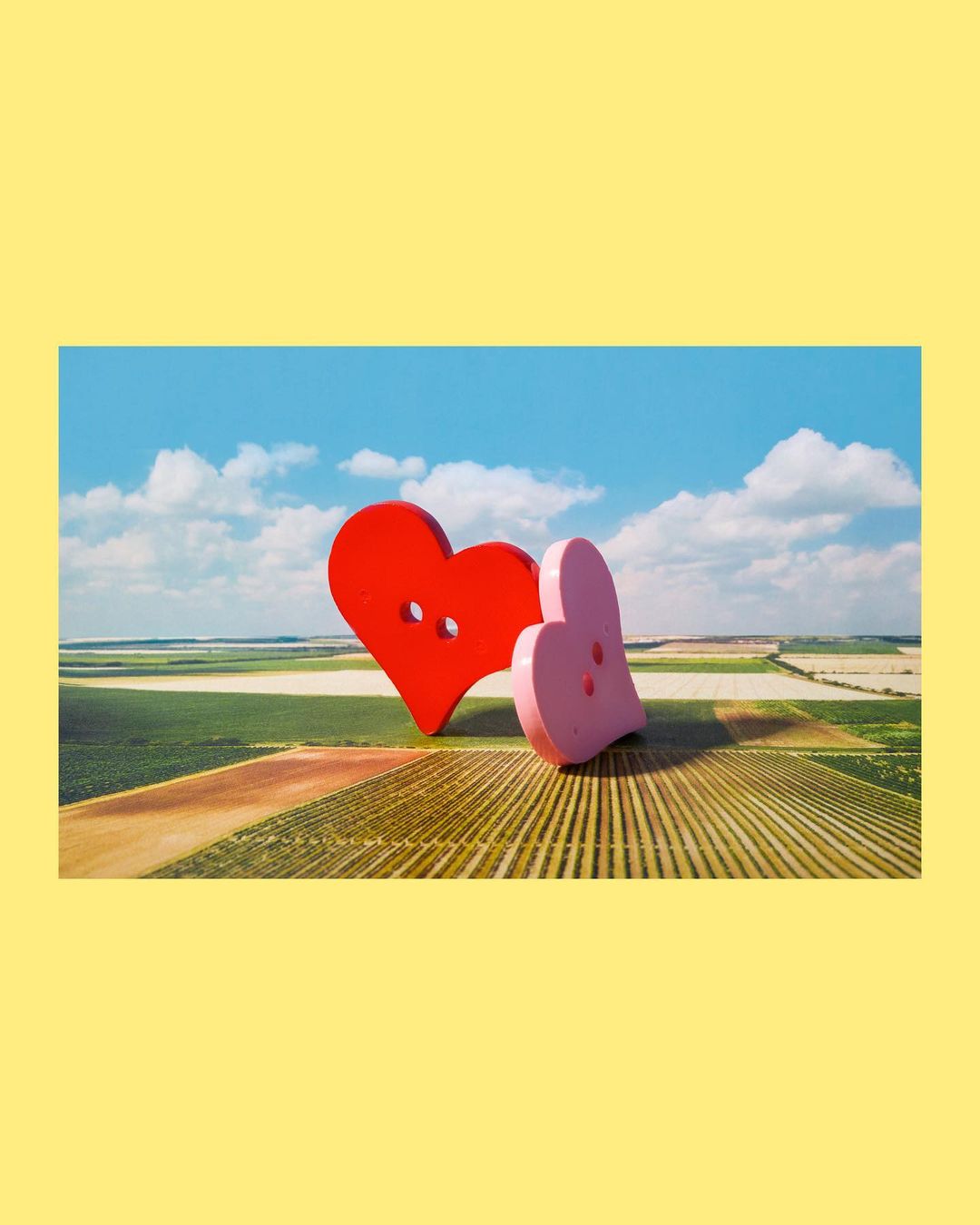 Two heart-shaped buttons, one red and one pink, sit on a striped surface with a backdrop of blue sky and clouds in a whimsical scene.