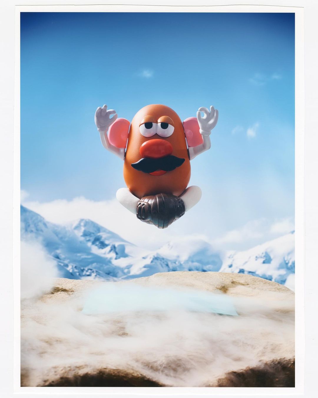 Mr. Potato Head levitates in a cross-legged pose above tan terrain with snowy mountains and a blue sky as a backdrop, resembling yoga.