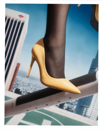 Close-up of a glossy yellow stiletto high heel shoe and black hosiery against a backdrop of a city skyline from a rooftop view.