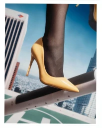 Close-up of a glossy yellow stiletto high heel shoe and black hosiery against a backdrop of a city skyline from a rooftop view.