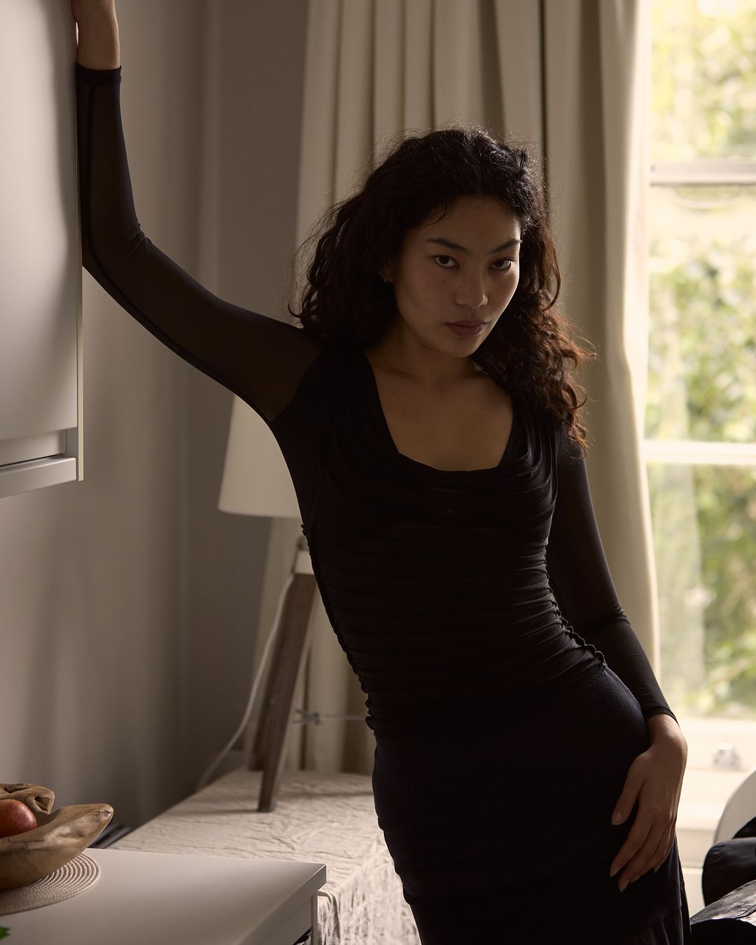 Woman with long dark curly hair wearing a fitted black long-sleeve top leaning against a wall indoors by a window