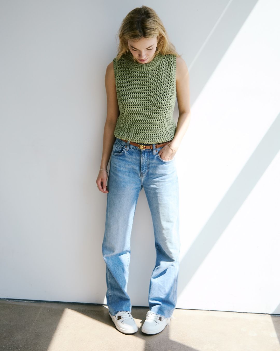 A young woman poses in a light green knit tank top and jeans, illuminated by natural light casting shadows on a white background.