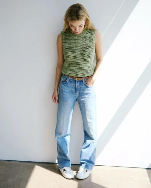 A young woman poses in a light green knit tank top and jeans, illuminated by natural light casting shadows on a white background.