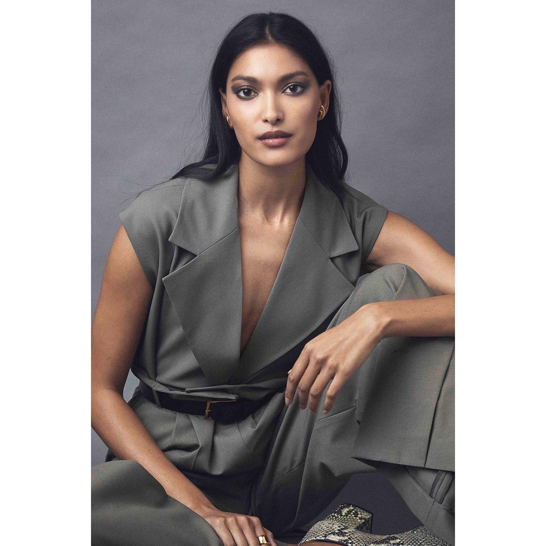 Elegant portrait of a South Asian woman in a green sleeveless suit, seated with a neutral expression against a solid grey backdrop.