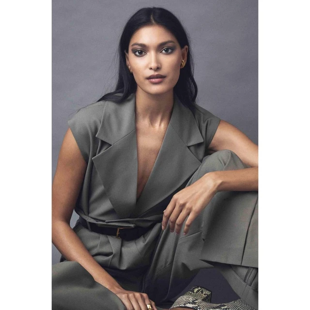 Elegant portrait of a South Asian woman in a green sleeveless suit, seated with a neutral expression against a solid grey backdrop.