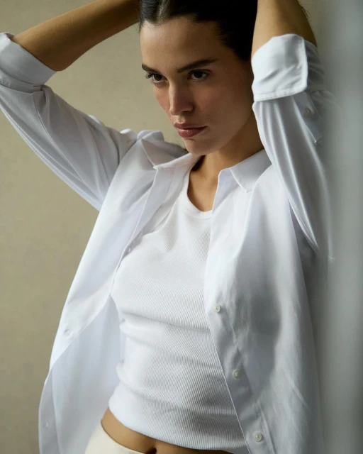 A young woman with her hands in her hair, wearing a white shirt, poses in an indoor setting with soft lighting.