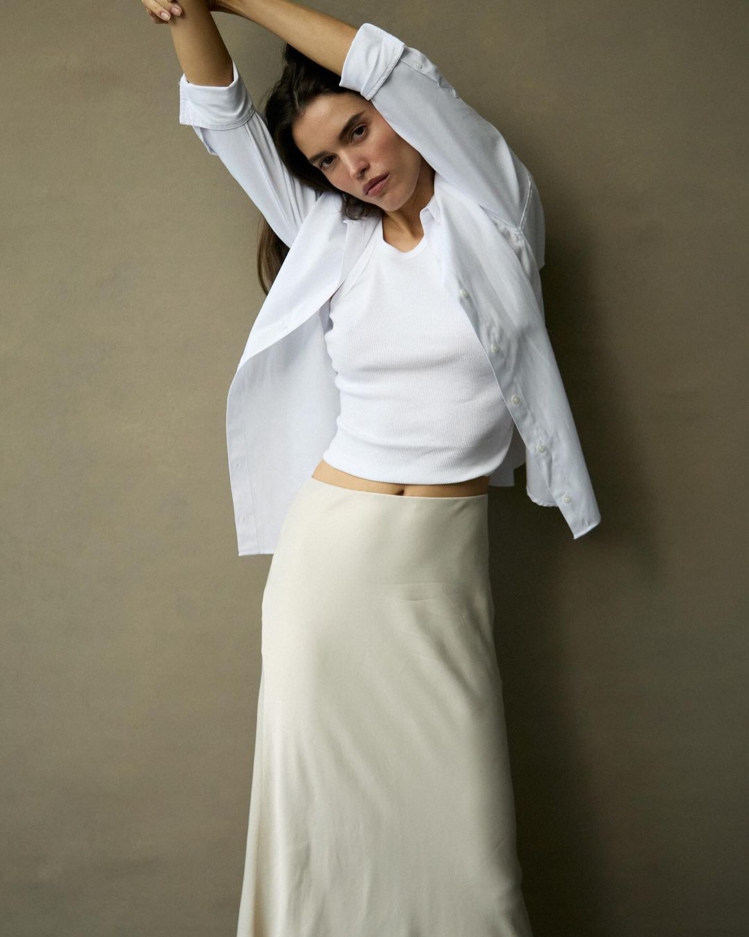 Woman in a white top and long skirt poses in front of a neutral background, creating a minimalist fashion portrait, studio lighting.