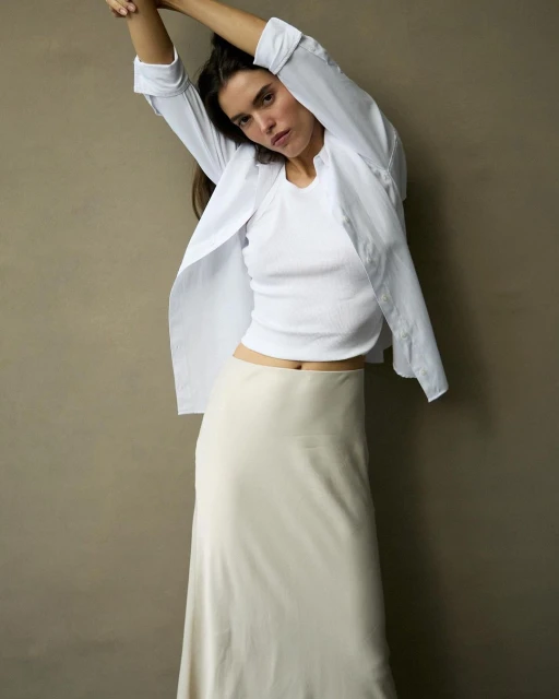 Woman in a white top and long skirt poses in front of a neutral background, creating a minimalist fashion portrait, studio lighting.