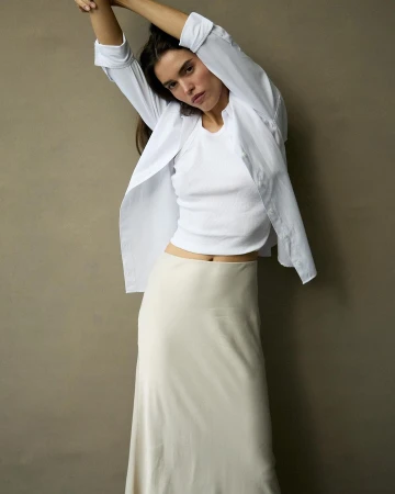 Woman in a white top and long skirt poses in front of a neutral background, creating a minimalist fashion portrait, studio lighting.