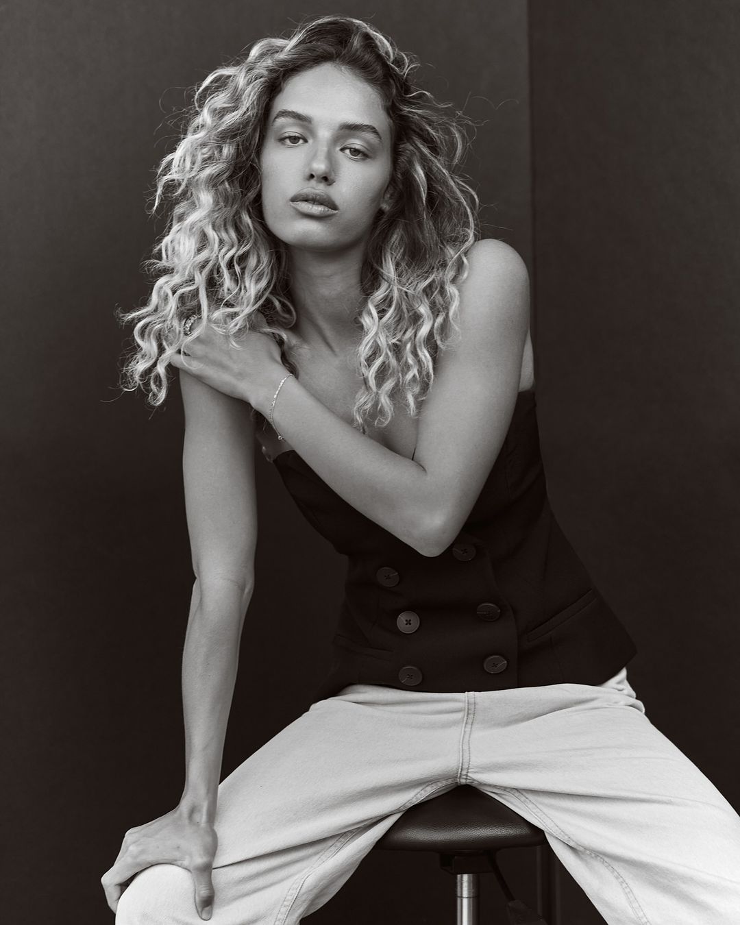 Portrait of a woman with curly blonde hair, wearing a black top and light jeans, sitting on a stool in front of a black backdrop.